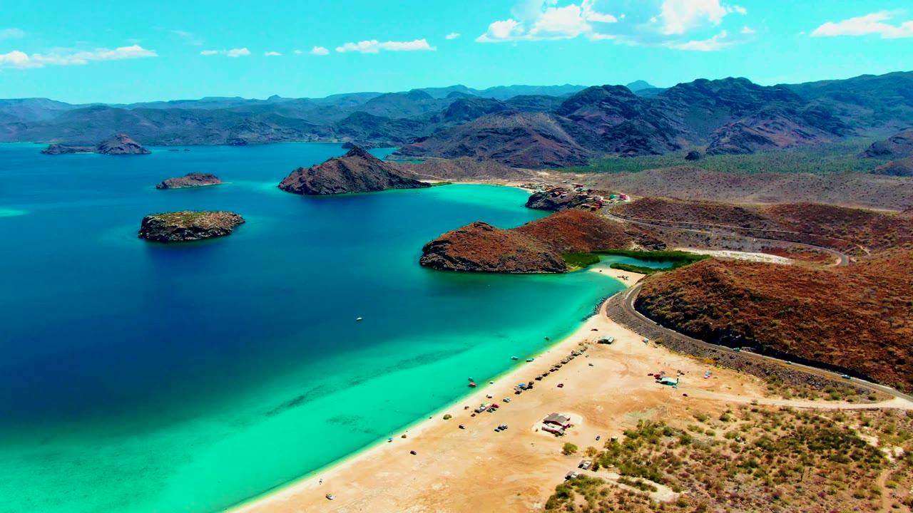 Third frame from the high aerial view at Playa Santispac, with the beach and road angled across the lower right, a vivid turquoise cove through the center, islands offshore, and rugged mountains on the horizon.