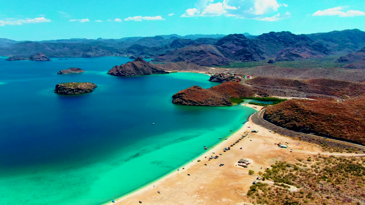 Aerial frame from Playa Santispac showing turquoise shallows along a sandy beach, darker bay water, offshore islands, desert hills, and tiny beach activity.