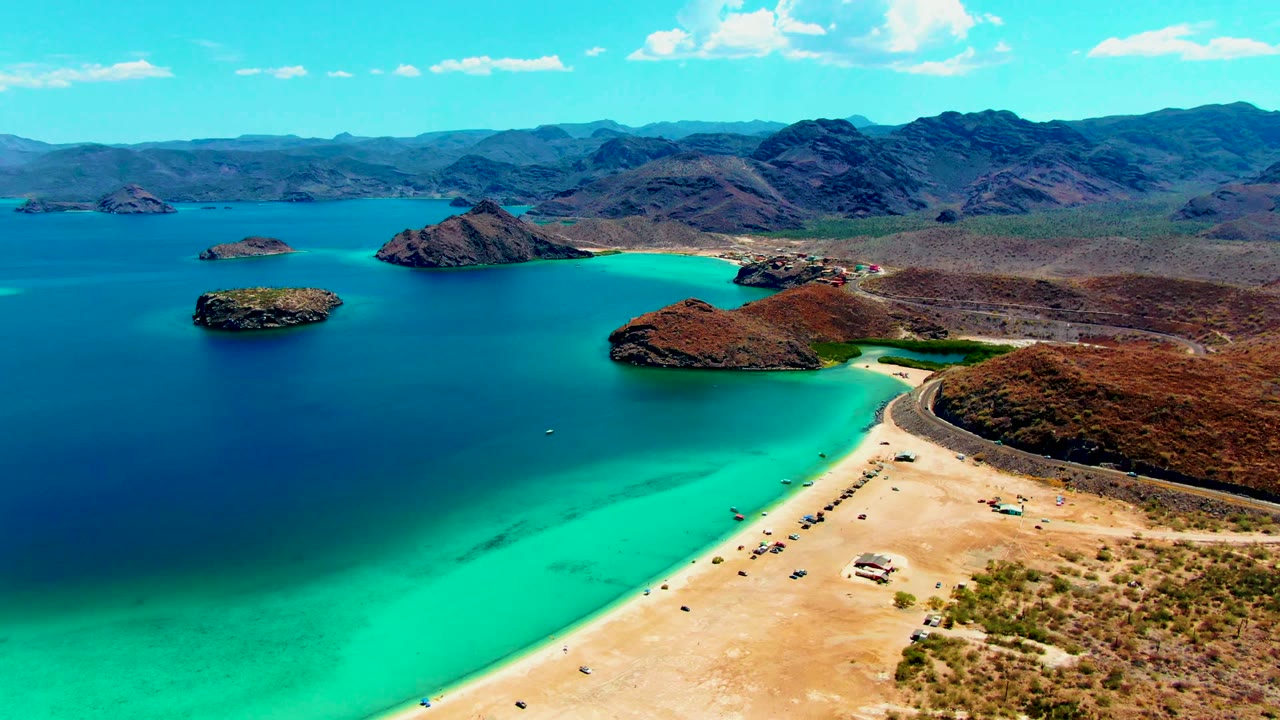 Wide aerial of sandy beach, turquoise water, islands, and mountain horizon.