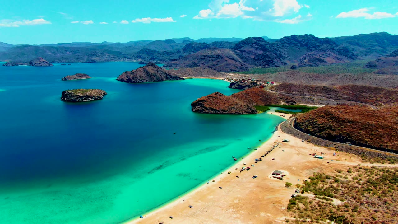 At 0:02, the Playa Santispac shoreline fills the lower frame with parked or moving vehicles on pale sand, clear turquoise water beside it, and islands backed by dry mountains.