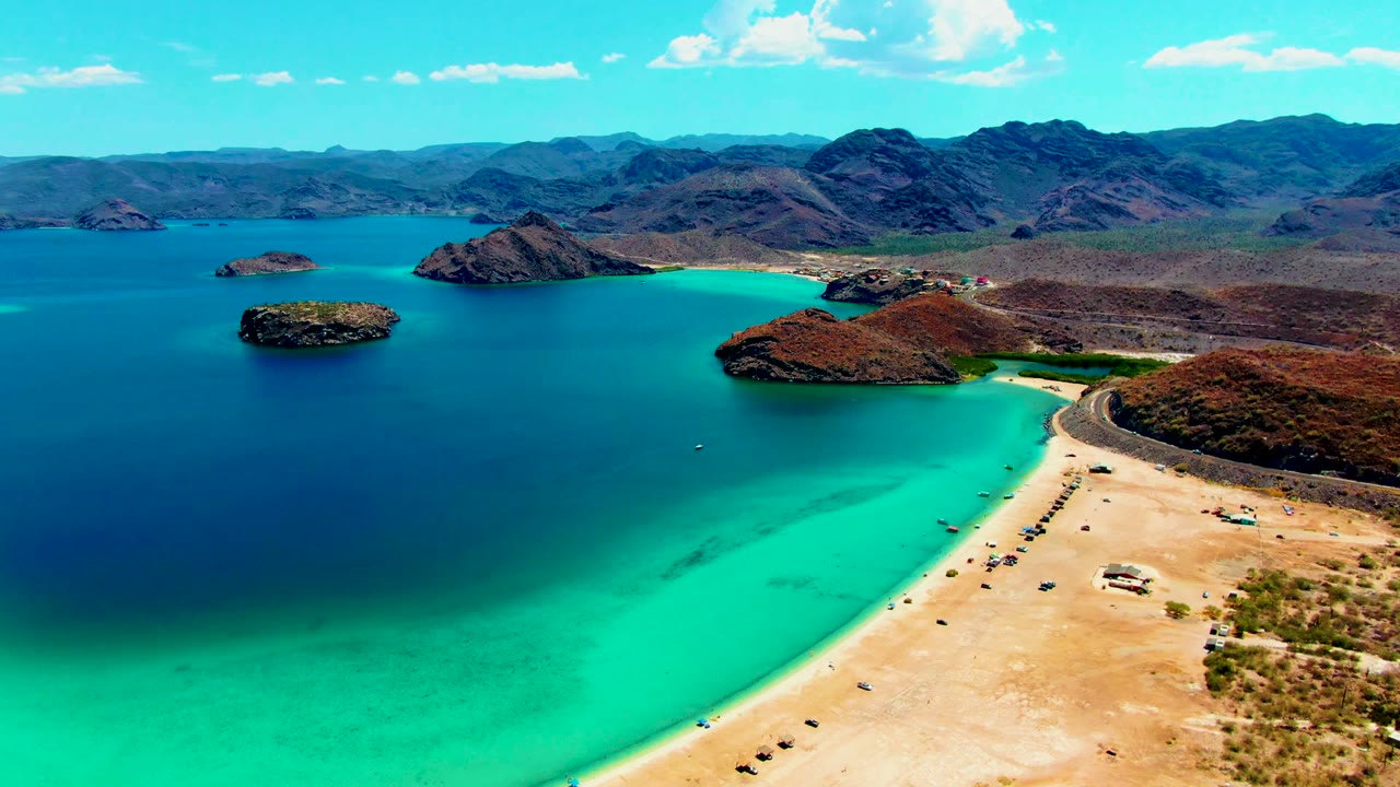 Aerial frame over Playa Santispac showing turquoise shallows along a sandy beach, darker blue bay water, rocky islands, desert hills, distant mountains, and tiny beachgoers near the shore.