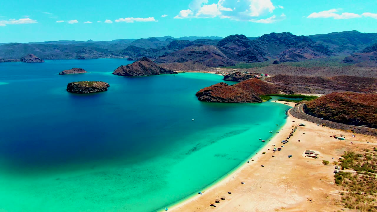 Wide aerial coastline with shallow turquoise water, sandy shore, rocky islands, and mountains.