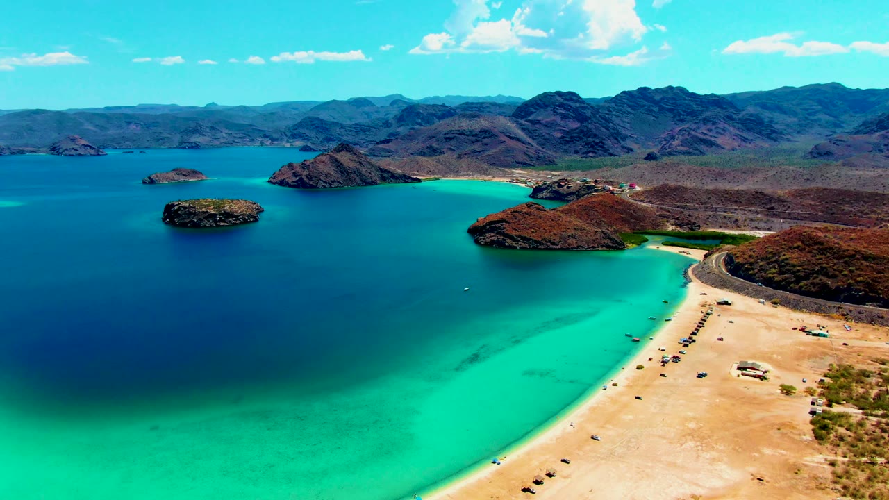 Aerial Playa Santispac view with the curved sand beach pushed to the lower right, bright turquoise shallows in the foreground, darker bay water to the left, rocky islands ahead, and distant mountains under scattered clouds.