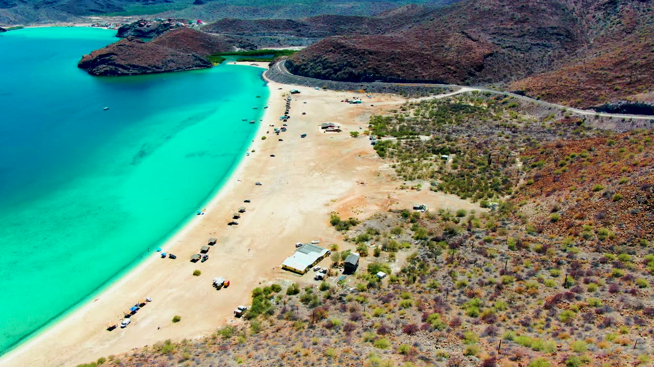 Aerial frame of Playa Santispac showing the turquoise bay on the left, a pale sandy beach with small vehicles, scrub vegetation to the right, and no visible people.