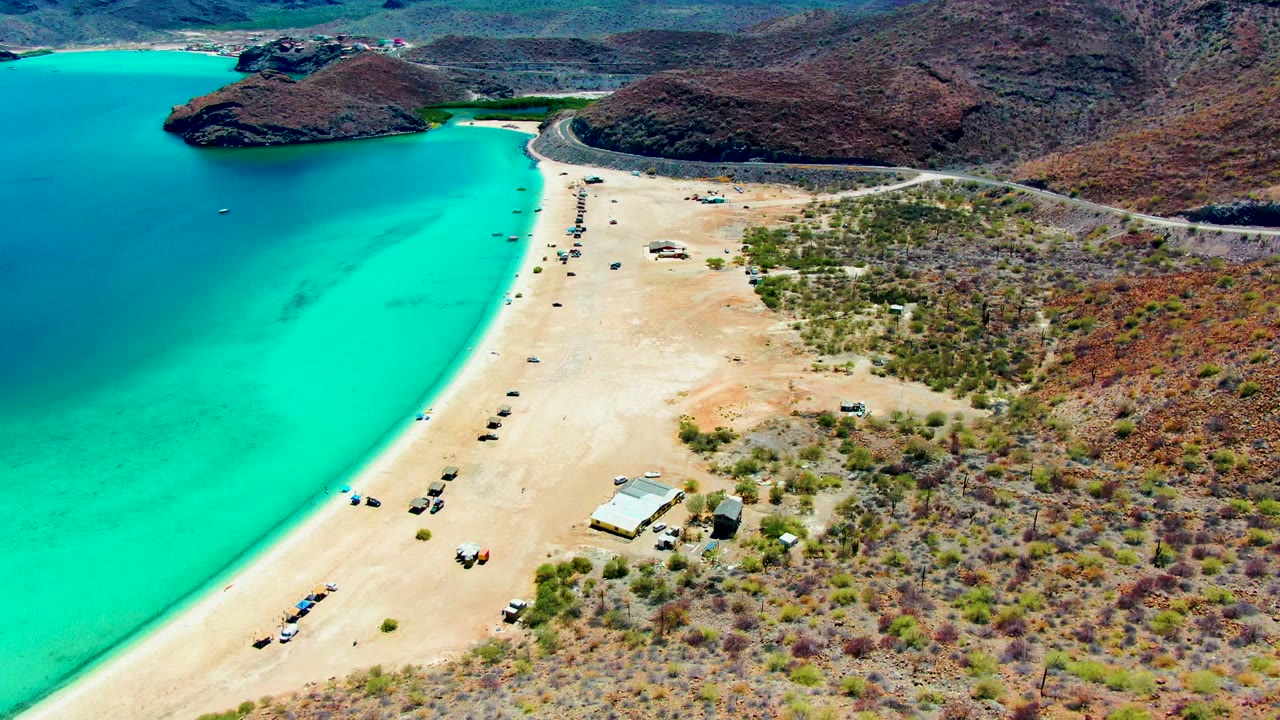 Aerial shoreline with bright turquoise water, sandy vehicle tracks, and arid hills.