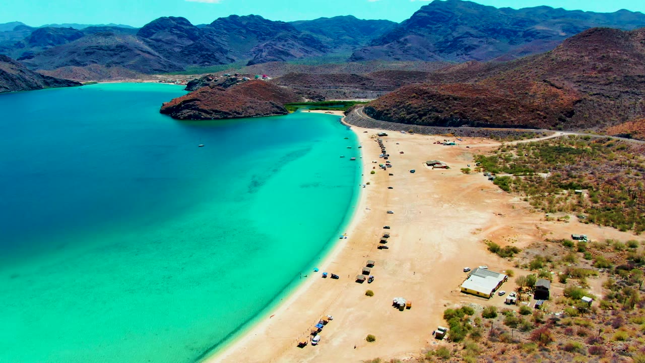 Aerial frame over Playa Santispac showing turquoise water on the left, a curving sandy beach with parked vehicles, arid scrubland, and mountains beyond, with no visible people.