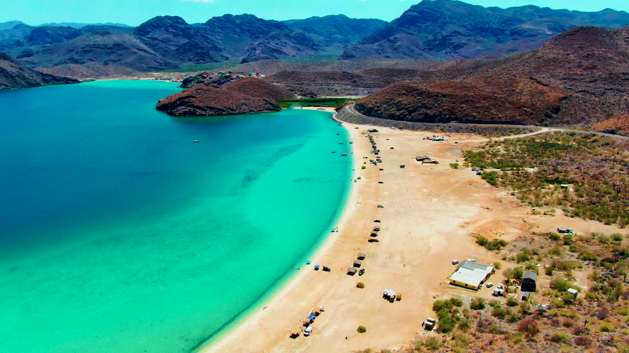 Wide aerial beach curve with turquoise water, vehicles on sand, and mountains behind.
