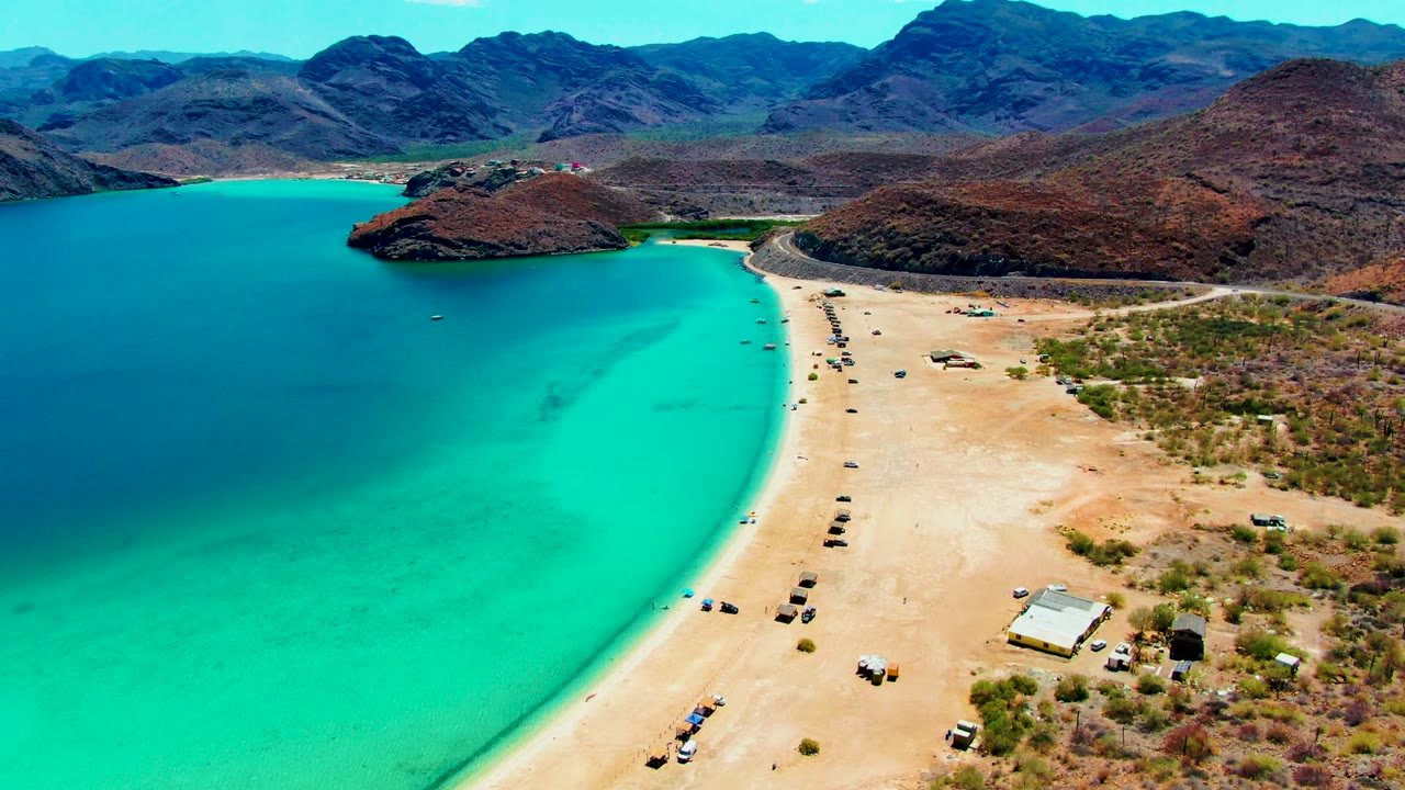 At 0:02, the aerial view shows Playa Santispac with the turquoise bay widening to the left, vehicles dotted along the sand, dry brown hills to the right, and no visible people.