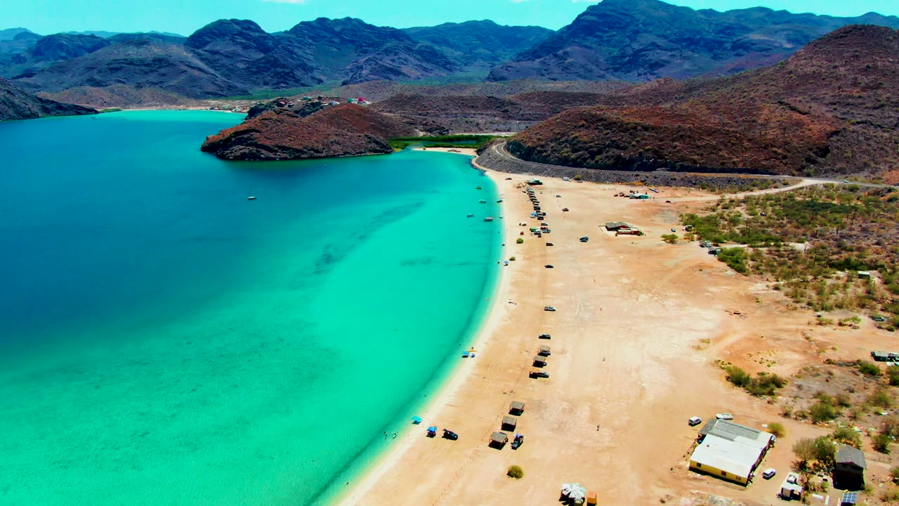 Opening aerial frame over Playa Santispac, with turquoise ocean on the left, a sandy beach with vehicles on the right, arid hills beyond, and no visible people.