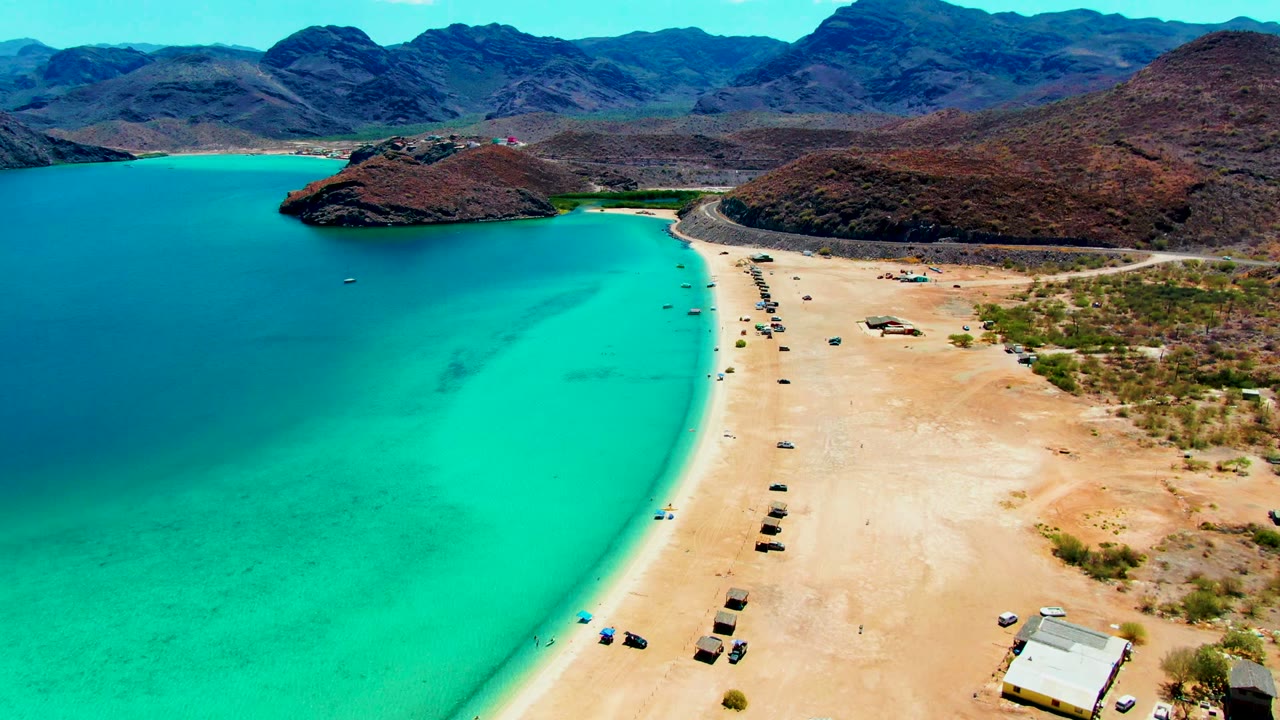Curving sandy shoreline with clear turquoise water and vehicles on the beach.