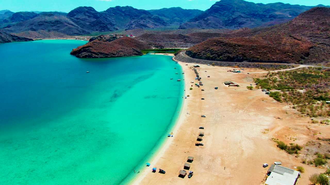 Mid-clip aerial frame of Playa Santispac with the turquoise bay occupying the left half, pale sand and vehicles on the right, dark mountains behind, and no visible people.