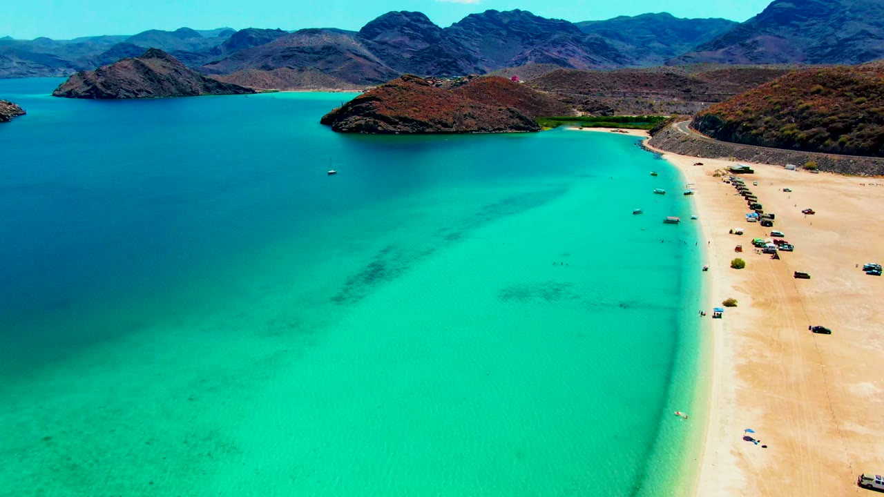 Aerial view over Playa Santispac, Baja California Sur, with turquoise water in the foreground, a sandy beach with vehicles on the right, and rugged hills across the bay.