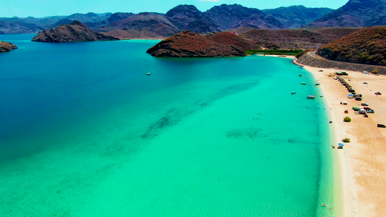 Aerial view of Playa Santispac with shallow turquoise water filling the lower frame, a narrow sandy beach on the right, sparse vehicles, and brown coastal hills behind the bay.