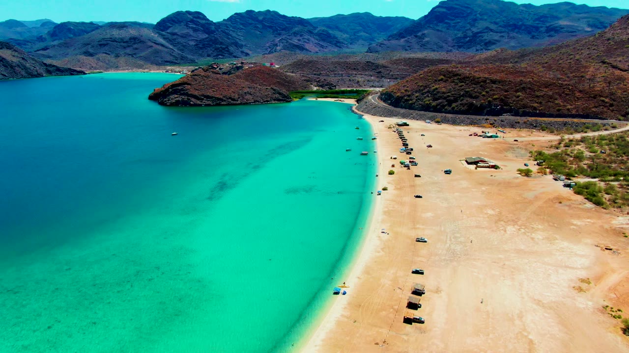 Aerial frame over Playa Santispac showing turquoise water on the left, a pale sandy beach with scattered vehicles on the right, rocky hills in back, and no visible people.