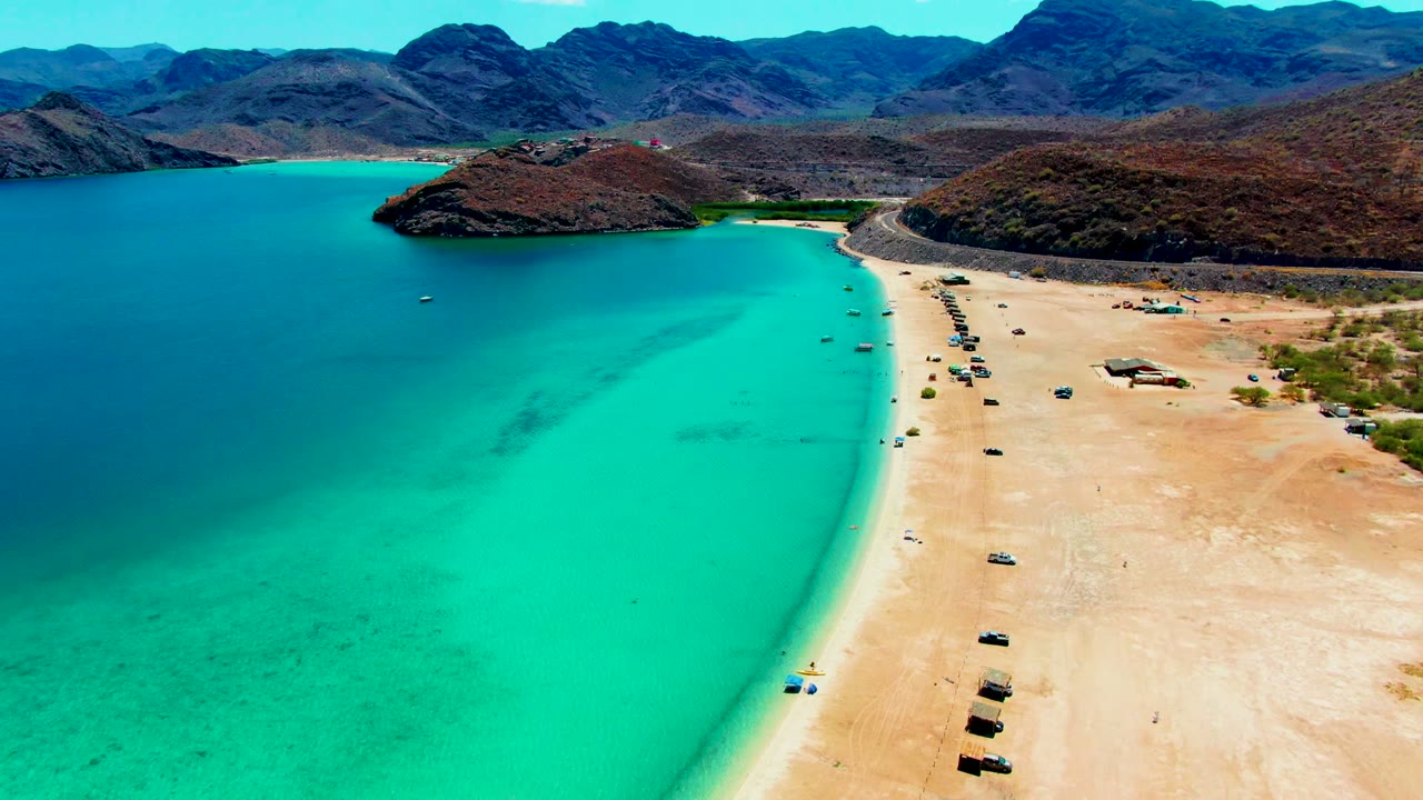 Wide drone view of clear water beside a vehicle-lined sandy shore.