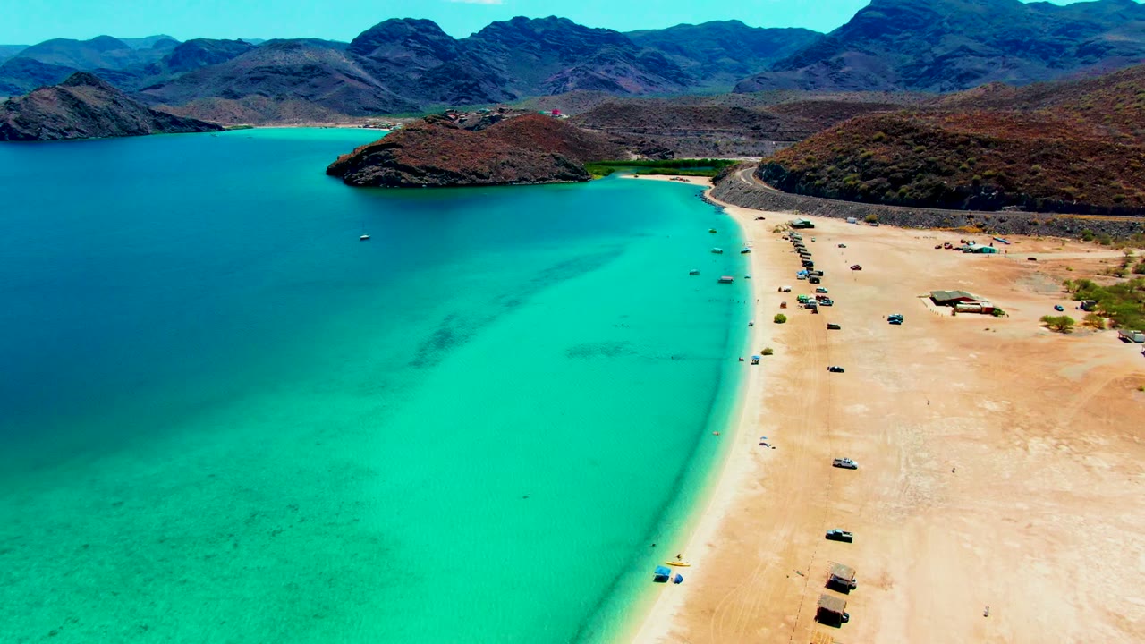 Aerial footage at Playa Santispac shows the shoreline running diagonally through frame, bright turquoise shallows, multiple small vehicles on sand, distant rocky mountains, and no visible people.