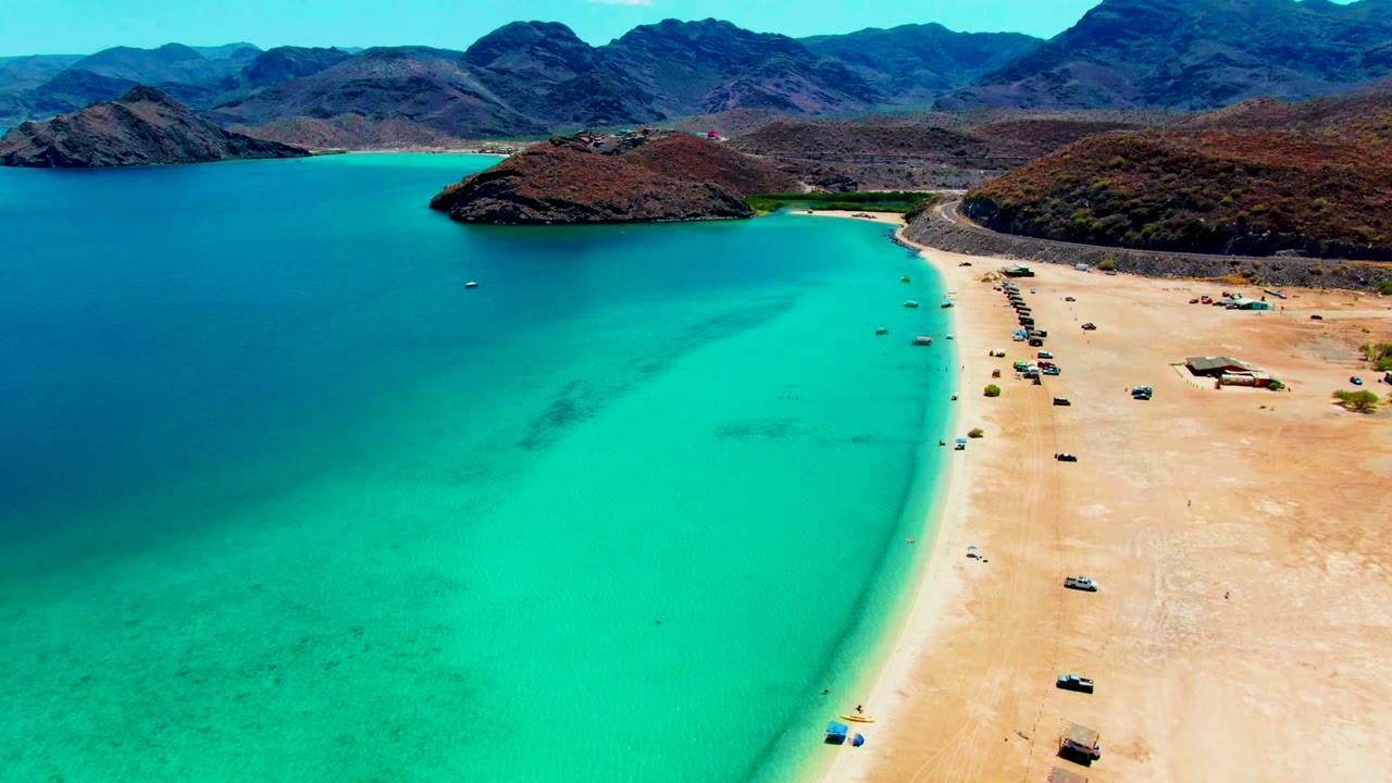 Aerial coastal view with deep blue water, turquoise shallows, beach vehicles, and hills.