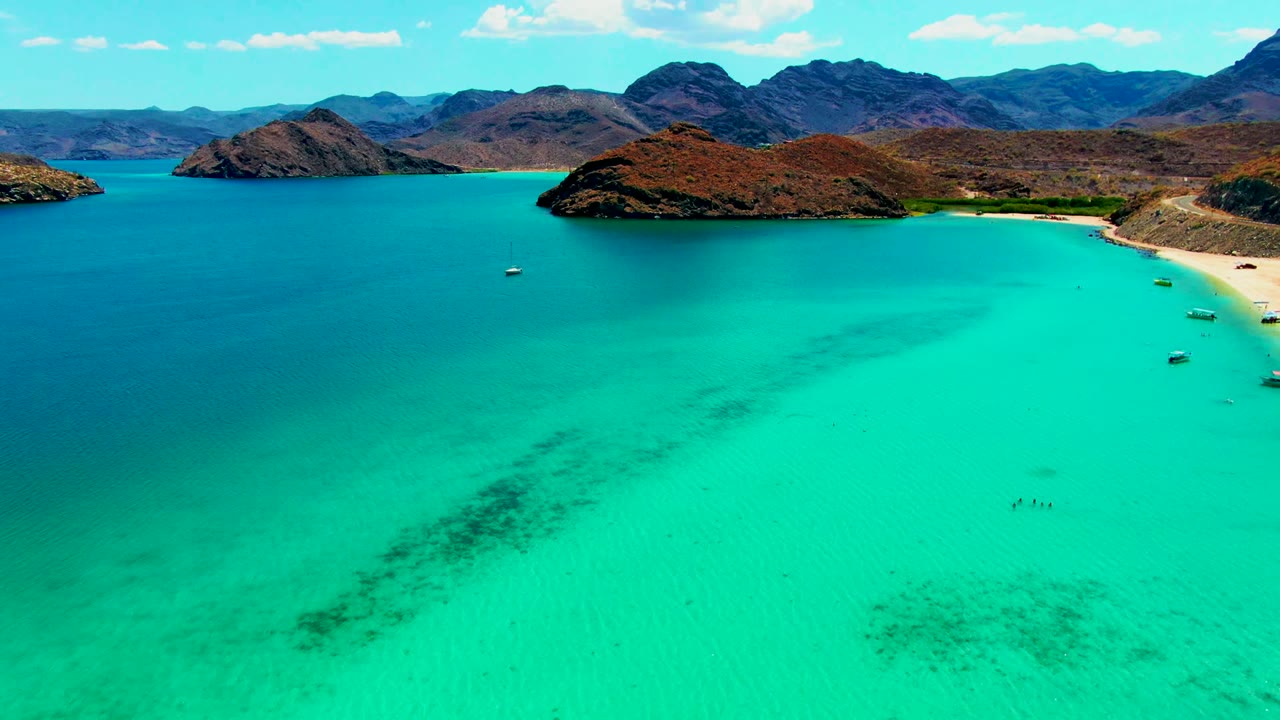 At 0:02, Playa Santispac appears from above with a large field of turquoise water, darker underwater patches, a curved sandy beach at right, small boats near shore, arid hills, distant mountains, and tiny swimmers visible.