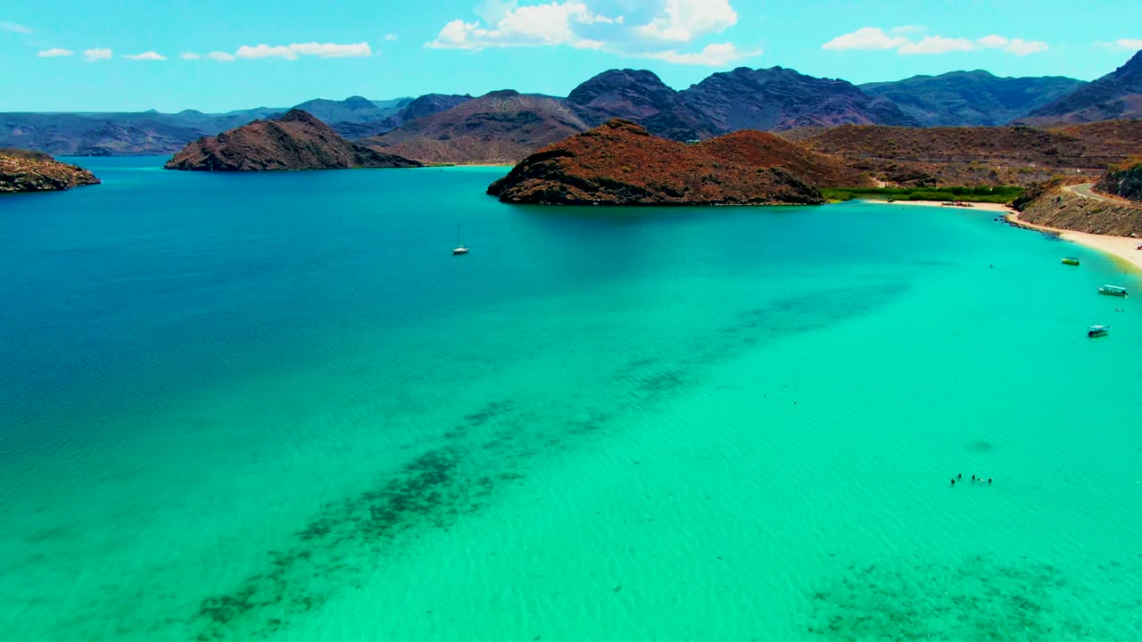 Aerial bay view with turquoise water, boats near the shore, swimmers, hills, and clouds.
