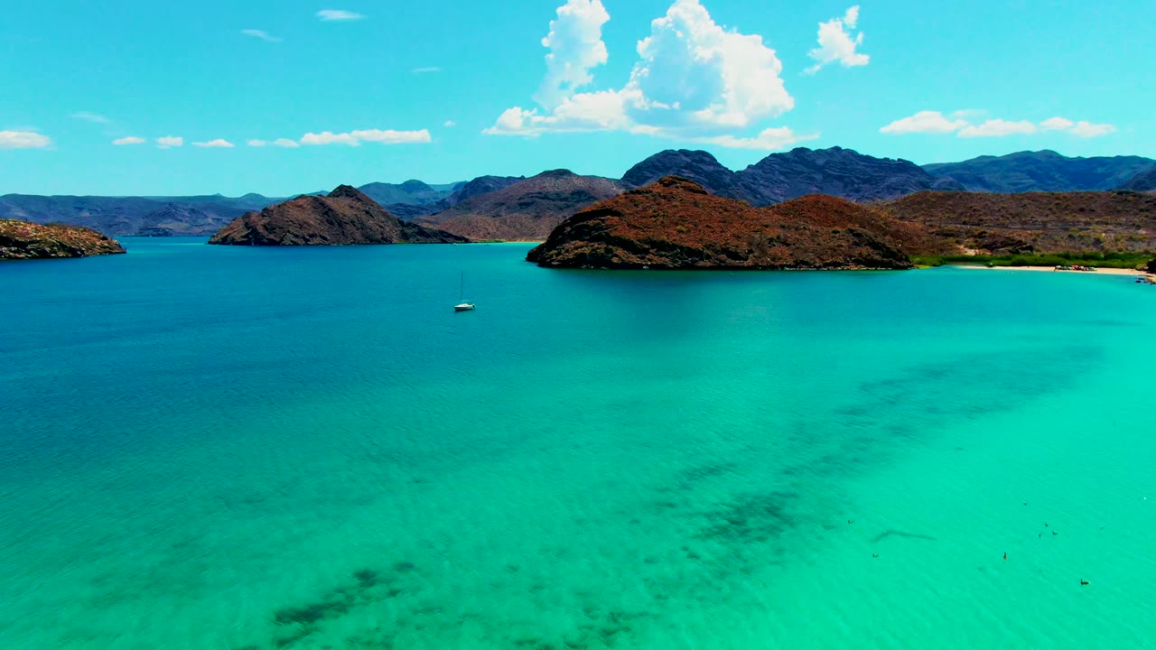 Aerial view over turquoise water at Playa Santispac, Baja California Sur, with a small sailboat left of center, rocky islands on the horizon, and no visible people.