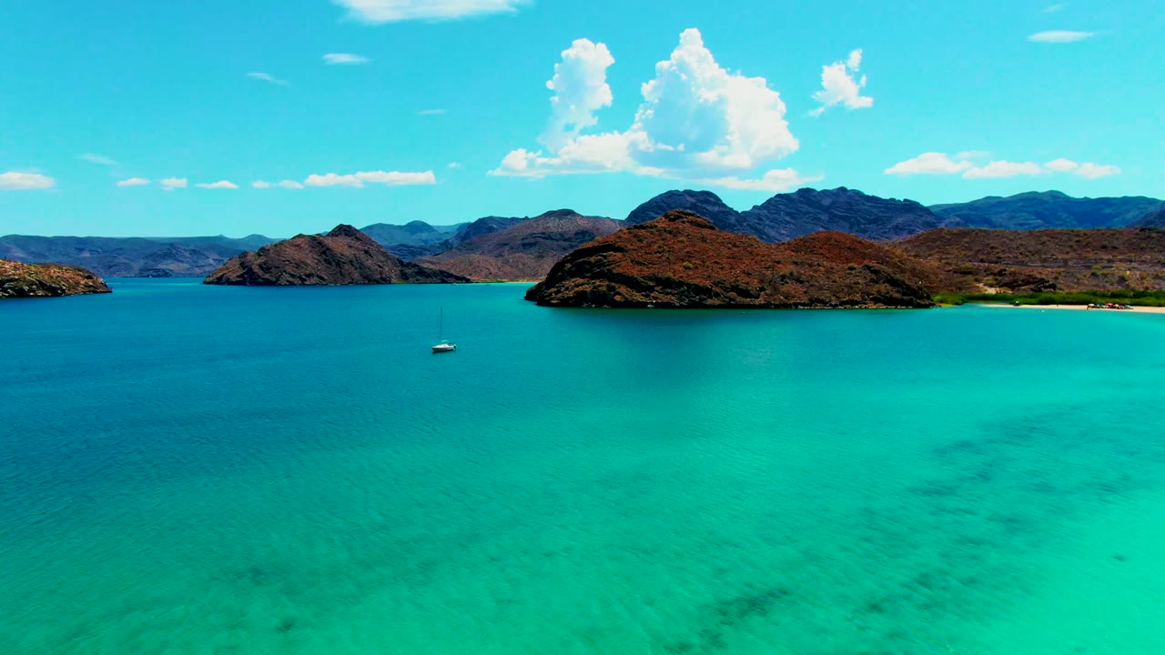 Wide aerial view of Playa Santispac water with the sailboat near the center-left, dark patches under the turquoise surface, rocky islands beyond, and no visible people.