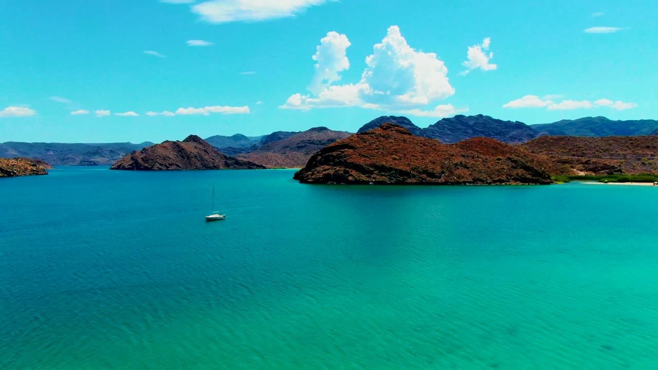 At 0:00, a white sailboat appears low on turquoise water at Playa Santispac, with rocky islands, distant ridges, bright clouds, and no visible people.