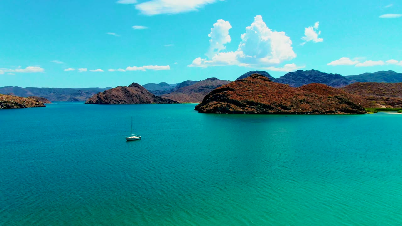 At 0:02, a small sailboat sits in open turquoise water beneath layered rocky islands and scattered white clouds at Playa Santispac, with no visible people.