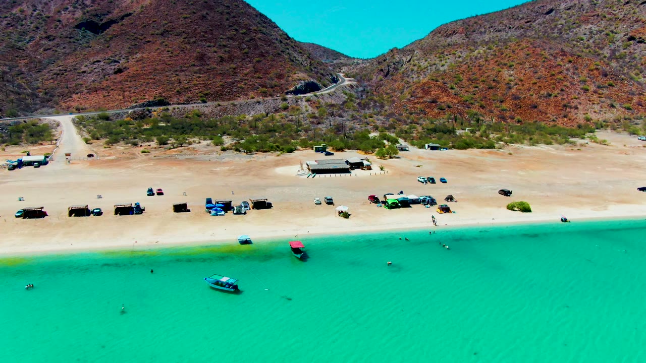 Aerial frame of Playa Santispac with turquoise water at the bottom, a small boat near shore, parked vehicles on the sand, dry mountain slopes behind, and no visible people.