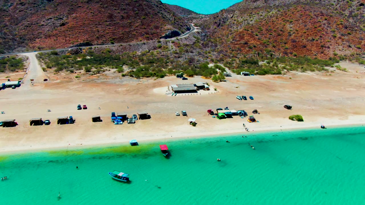 Mid-clip aerial frame at Playa Santispac with bright turquoise shallows in front, scattered vehicles and beach structures on the sand, mountain slopes in back, and no visible people.