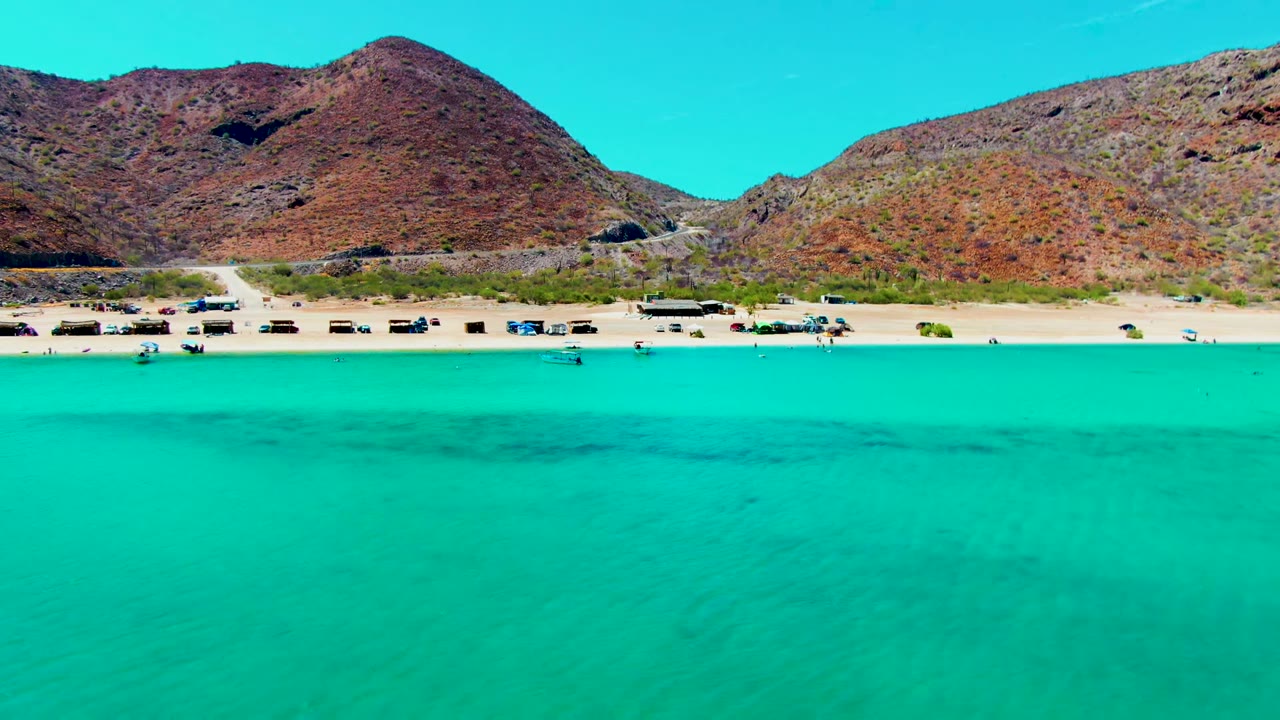 Opening aerial frame of Playa Santispac with broad turquoise water in front, a narrow sandy beach with parked vehicles, and reddish rocky hills across the background.