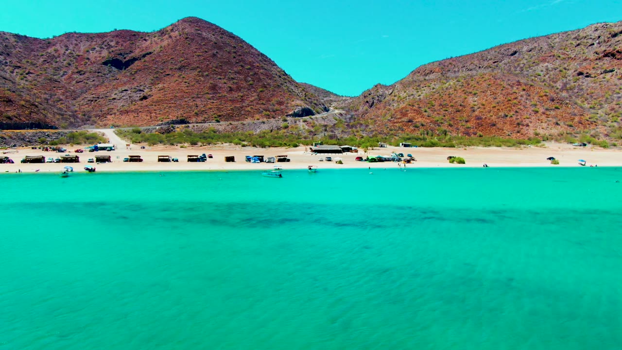 Aerial beach view with clear shallow water and a road below rocky hills.