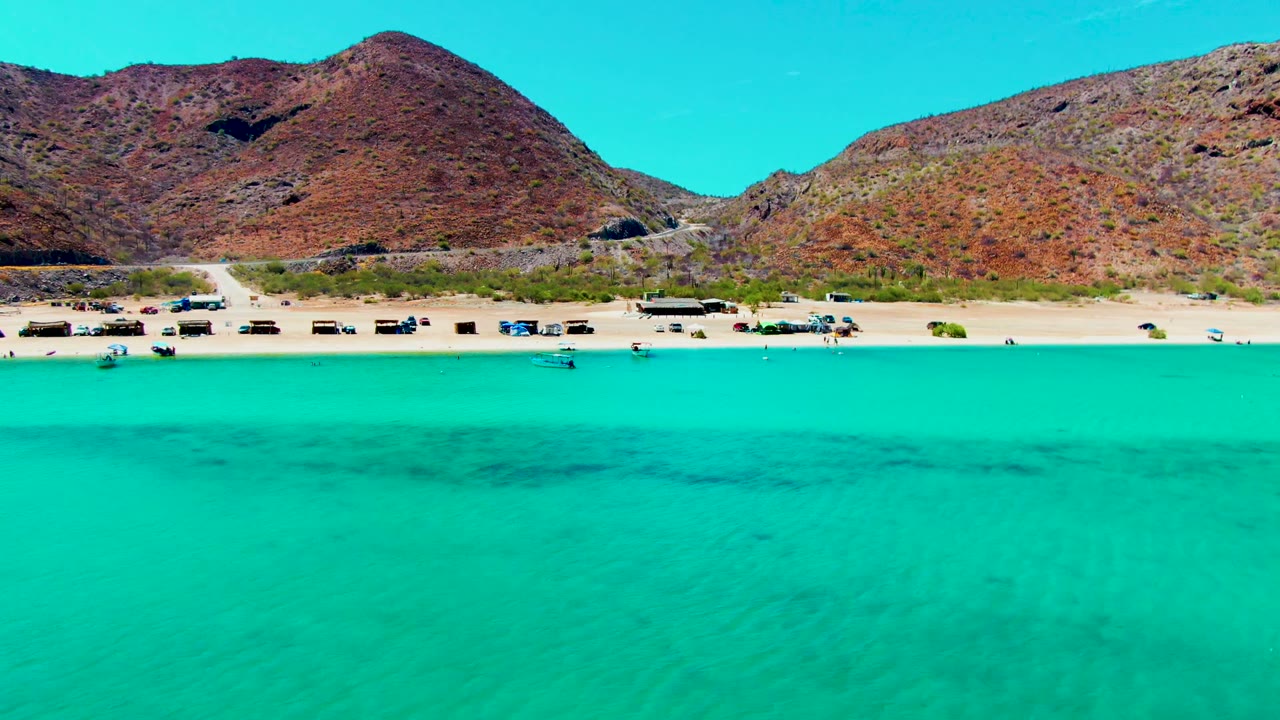 The aerial view holds on Playa Santispac with saturated turquoise water in the foreground, vehicles spaced along the sand, a low beach structure, and rugged hills behind.
