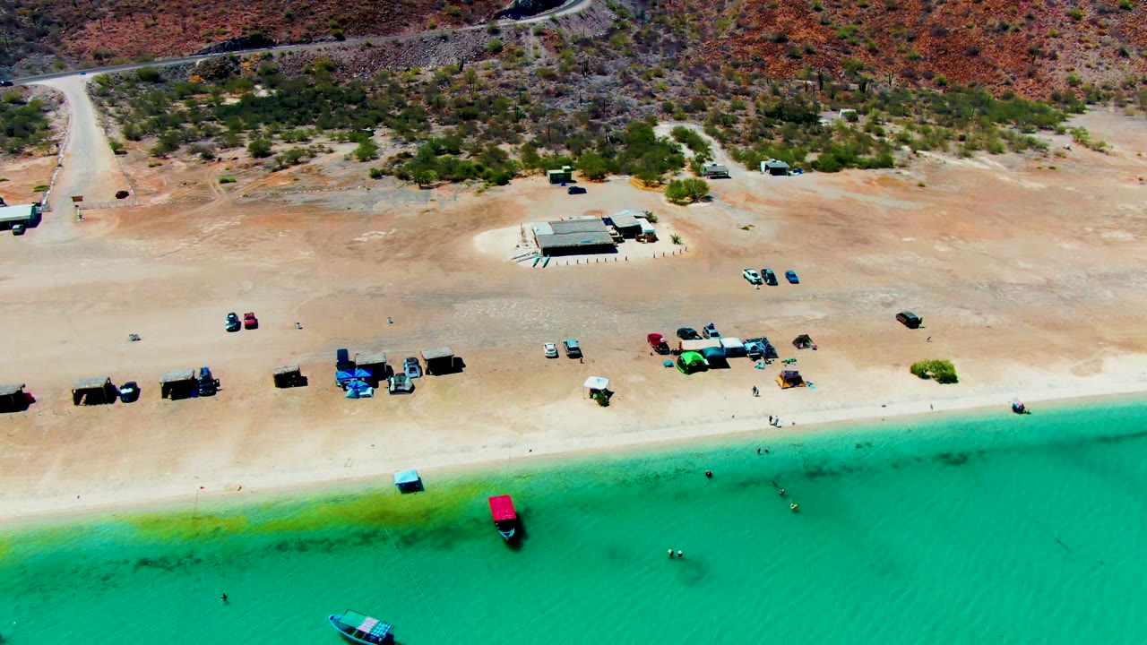Aerial frame at Playa Santispac showing turquoise water below a sandy beach with scattered vehicles, small boats, and dry hillside terrain in the background, with no visible people.