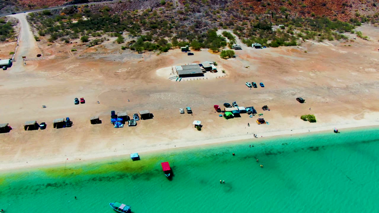 High aerial beach view with turquoise water, boats, vehicles, and dry hills.