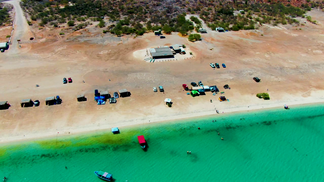 Aerial frame at Playa Santispac showing the turquoise shallows, sandy shoreline, parked vehicles, small boats near the water, and dry terrain beyond the beach, with no visible people.