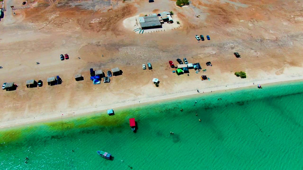 Overhead view of Playa Santispac with sandy shore, scattered parked vehicles, small shade structures, tiny swimmers in shallow turquoise water, and a greenish band near the beach.