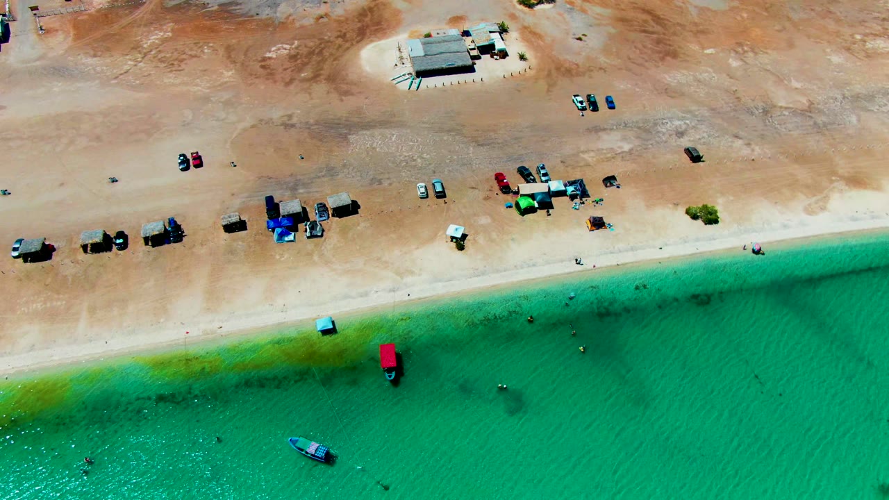 Aerial shoreline with parked cars, people in shallow water, and a small boat offshore.