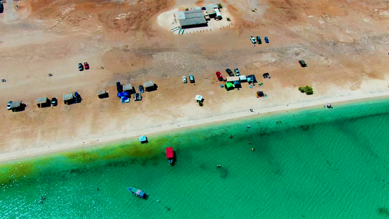 Drone-style overhead frame of Playa Santispac with clustered vehicles and tents along the sand, tiny swimmers near shore, and the green-blue water band shifted along the shallows.