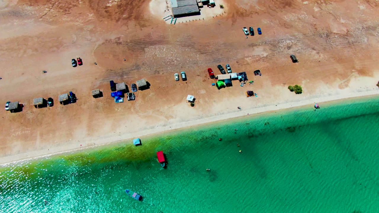 Aerial view over Playa Santispac with parked vehicles and small shelters on tan sand beside turquoise water, with no visible people at this scale.