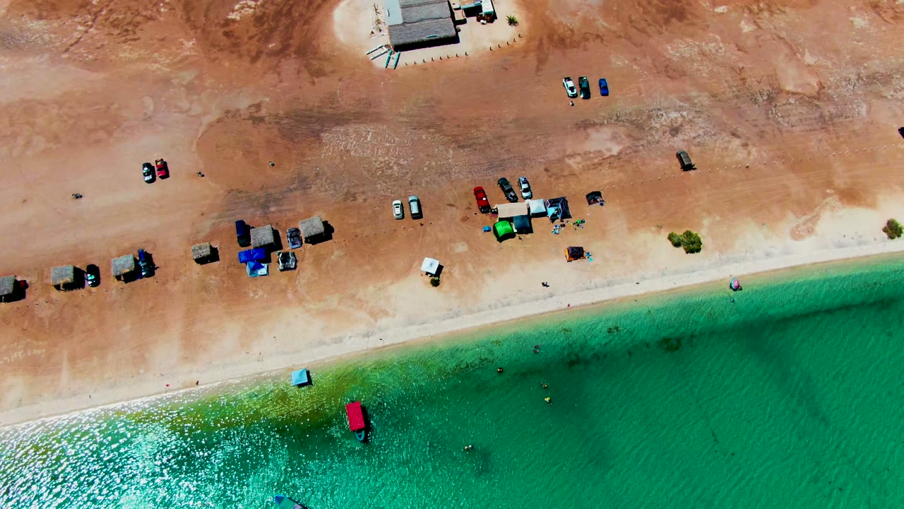 Aerial view of Playa Santispac with a broad sandy parking area, scattered vehicles, compact beach shelters, and turquoise-green water below, with no visible people at this scale.