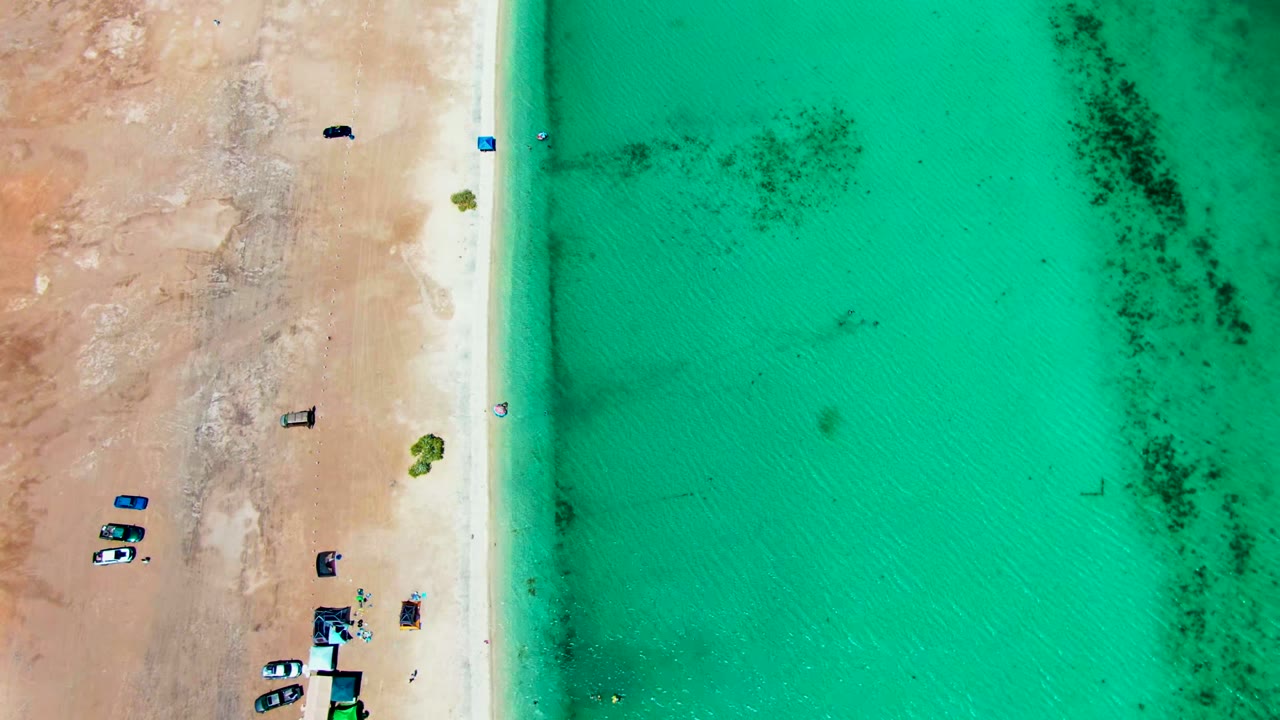 Overhead view of Playa Santispac with tan sand on the left, clear turquoise water on the right, several vehicles near the lower shore, and small beachgoers by the waterline.