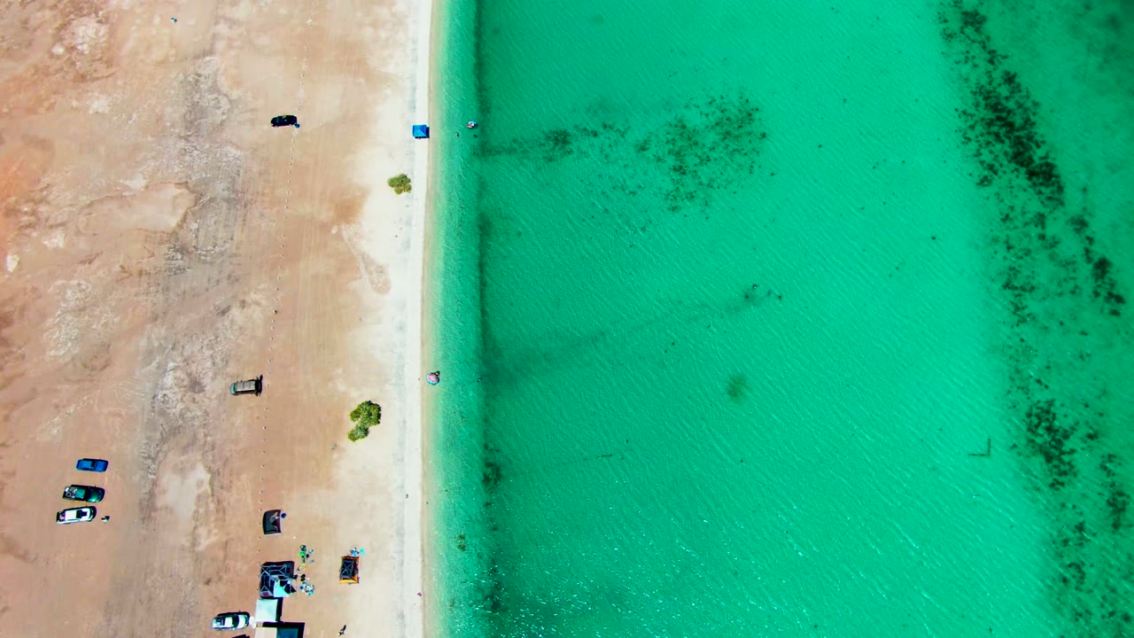 Top-down Playa Santispac frame with the sandy beach running vertically, vehicles and small beach setup lower left, scattered people near the water, and dark patches in the shallow turquoise water.