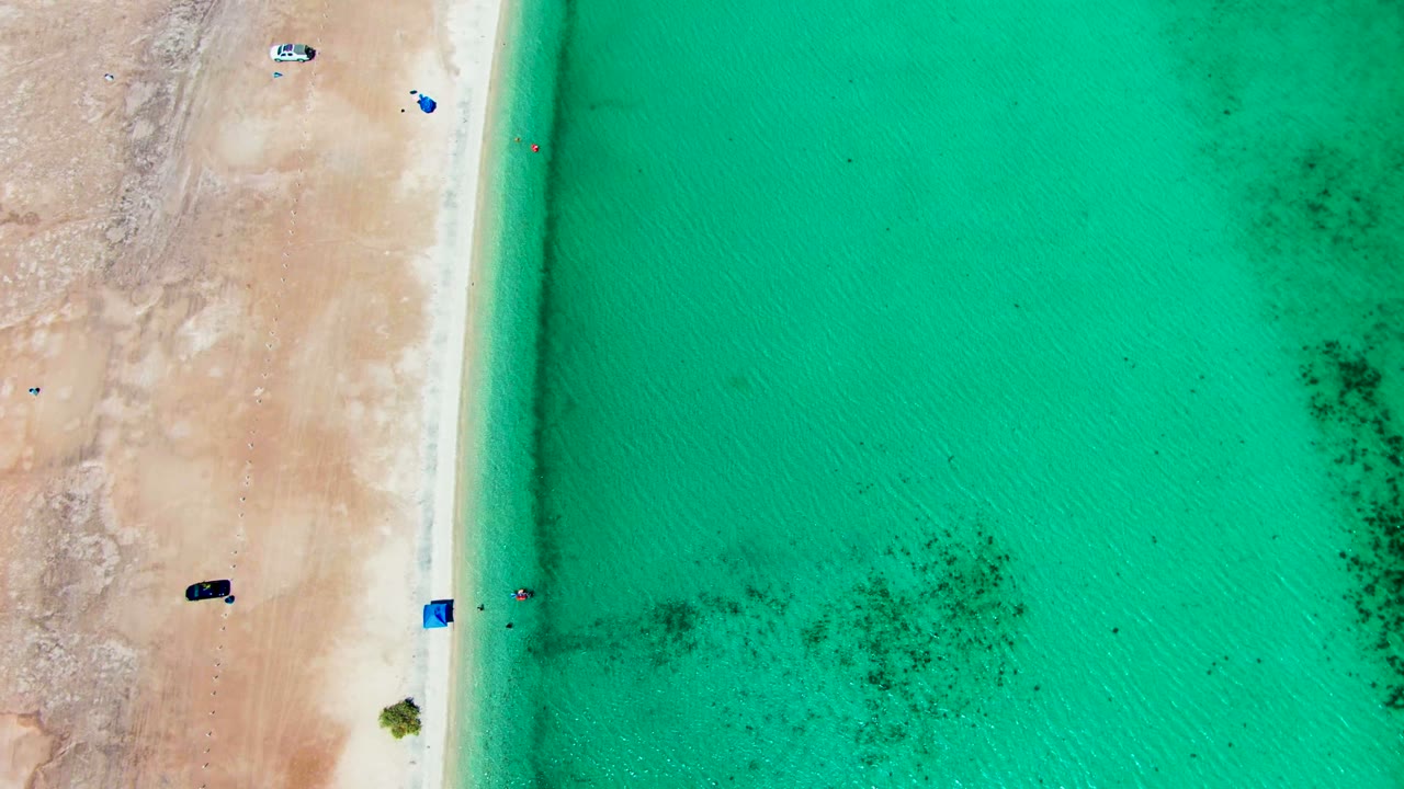 Aerial frame over Playa Santispac showing tan beach on the left, turquoise water on the right, several parked or moving vehicles, blue shade items, and tiny people near the shoreline.