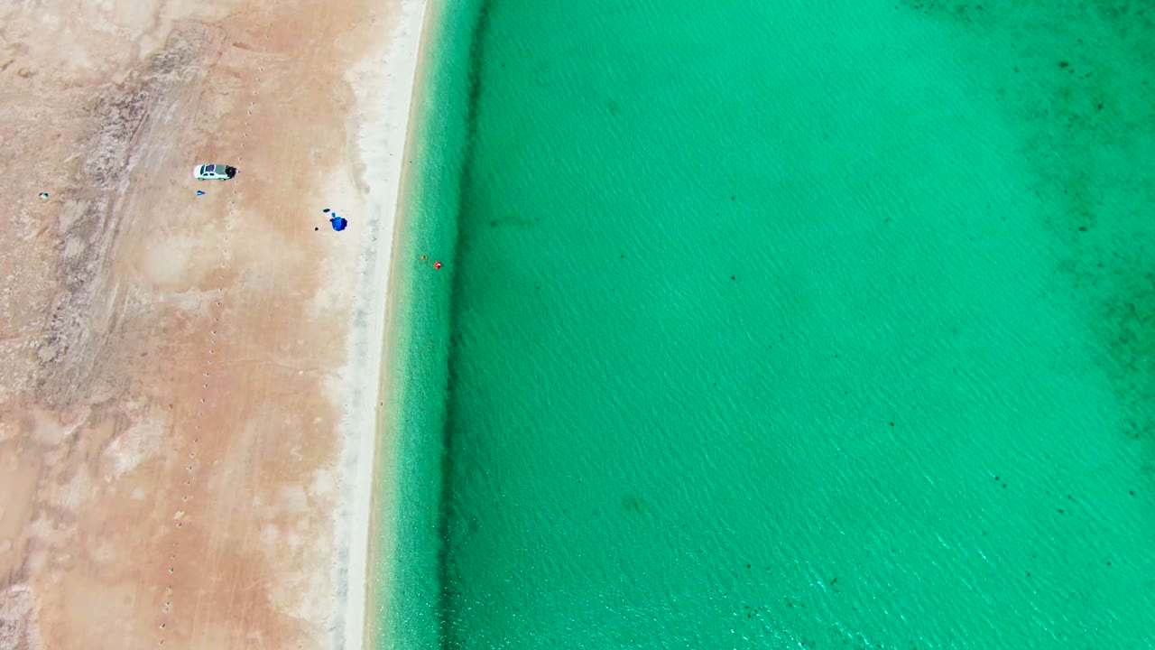 Playa Santispac appears from above with more open turquoise water filling the right side, a pale sand strip on the left, vehicles near the upper beach, and small colored beach items close to shore.