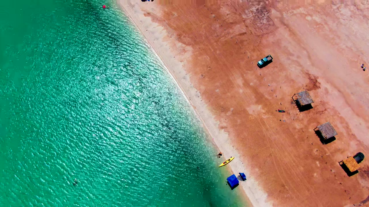 Top-down aerial frame at Playa Santispac showing rippled turquoise water on the left, a narrow pale shoreline, reddish sand, small vehicles, thatched shade structures, visible people, and a yellow kayak near the waterline.