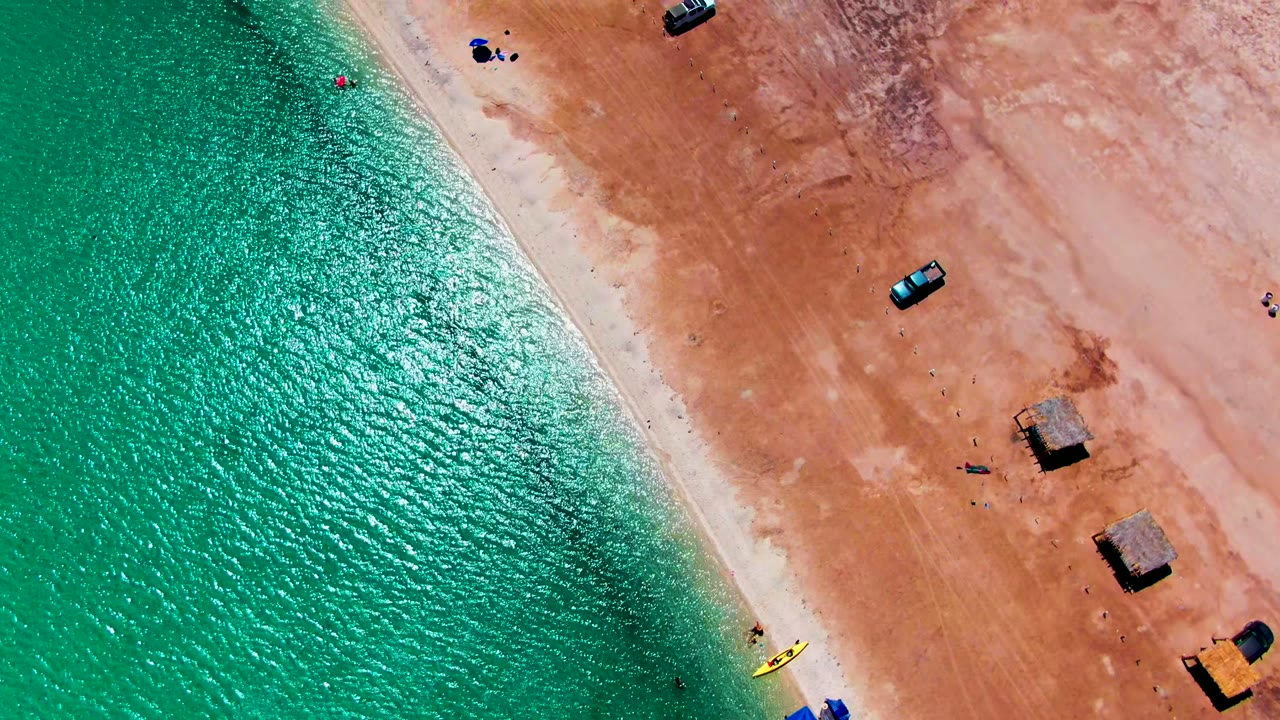 Overhead beach view with turquoise water, yellow kayak near shore, small vehicles, huts, and people on sand.