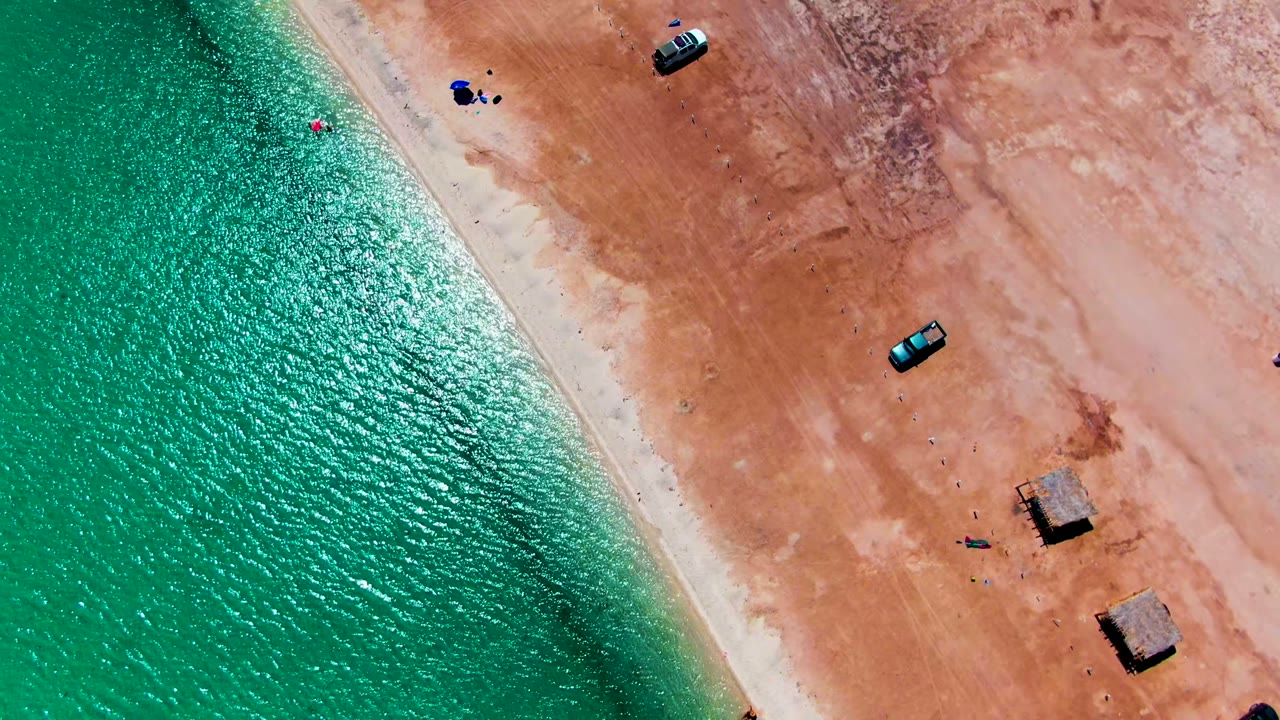 At Playa Santispac, the aerial view shifts to show more open reddish sand on the right, rippled turquoise water on the left, small vehicles near the upper and middle beach, visible people near blue shade, and thatched huts lower right.