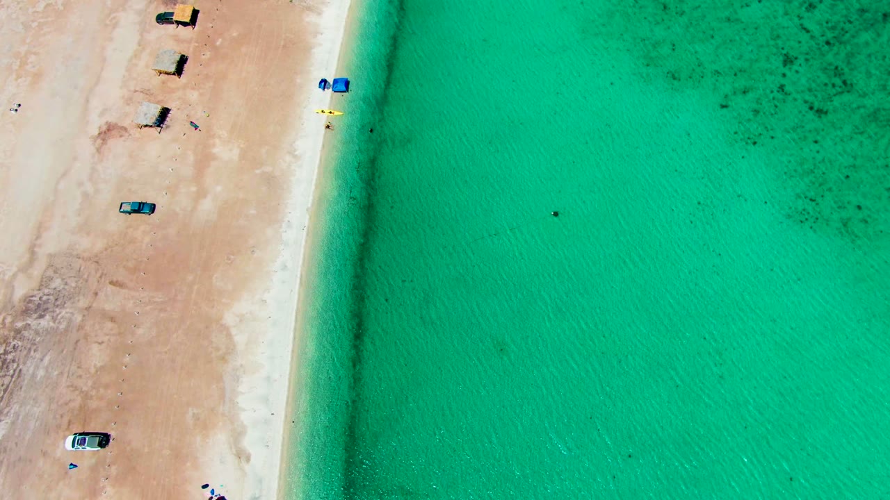 Overhead view of Playa Santispac with tan sand on the left, turquoise water on the right, several small vehicles and shade huts, and tiny swimmers near the shoreline.