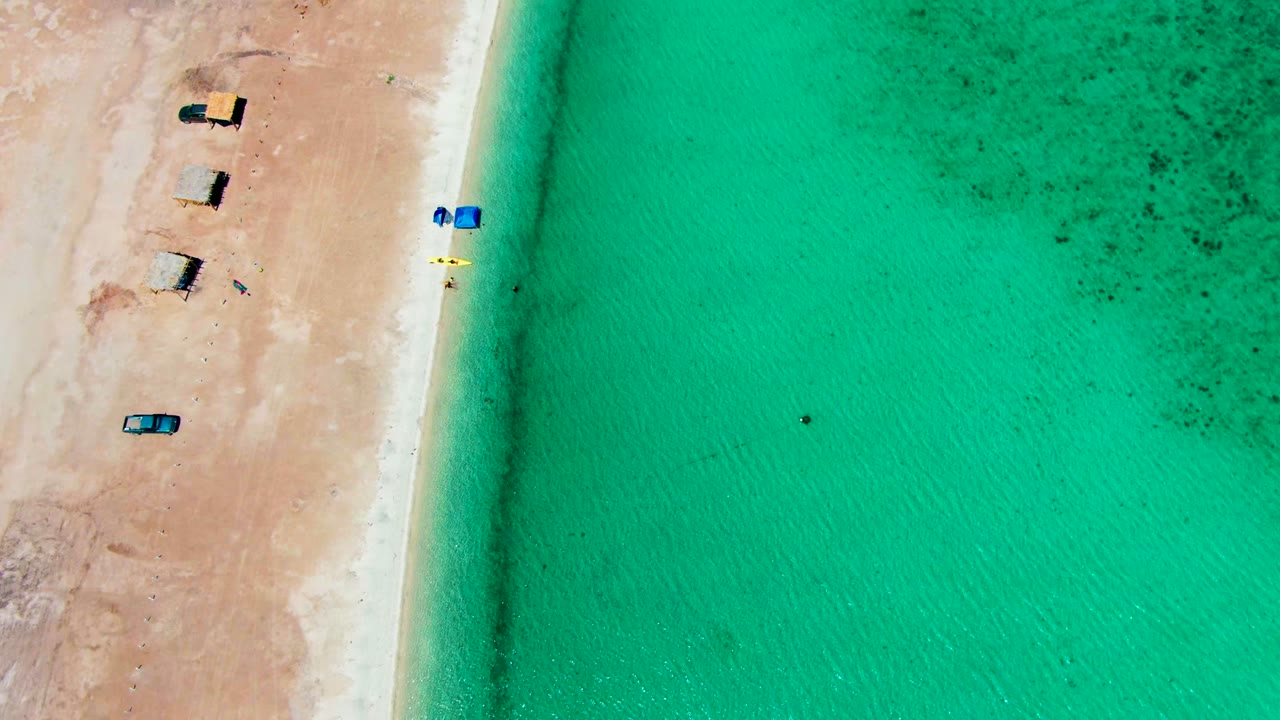 Aerial frame over Playa Santispac showing the beach edge shifted slightly, with thatched huts and vehicles on sand, blue canopy near the water, and small people visible in the turquoise shallows.