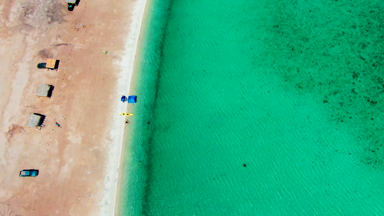 Top-down Playa Santispac frame with a narrow pale shoreline between dry sand and turquoise water, several huts and vehicles on the beach, and tiny swimmers offshore.